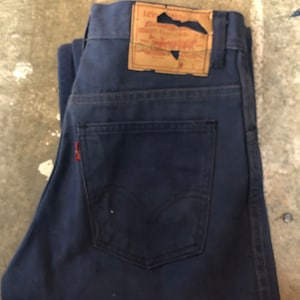 Può includere: Jeans di denim blu scuro con un classico design a cinque tasche. Presenta un'etichetta rossa Levi's sulla tasca posteriore e una toppa in pelle invecchiata. I jeans sono piegati, mostrando le tasche posteriori e il logo Levi's.