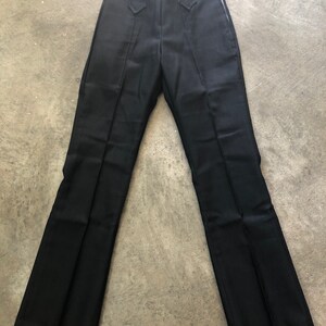 May include: Black dress pants with a high waist and a flared leg. The pants have a zipper closure and decorative stitching details. The fabric appears to be a smooth, woven material. The pants are laid flat on a concrete surface.