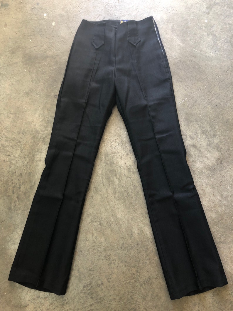 May include: Black dress pants with a high waist and a flared leg. The pants have a zipper closure and decorative stitching details. The fabric appears to be a smooth, woven material. The pants are laid flat on a concrete surface.