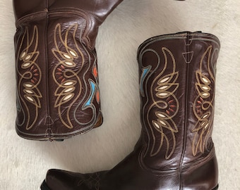 Könnte beinhalten: Ein Paar braune Leder-Cowboystiefel mit dekorativen Nähten in Gold, Orange, Weiß und Blau. Die Stiefel haben spitze Zehen und gestapelte Absätze und präsentieren einen klassischen Western-Stil.