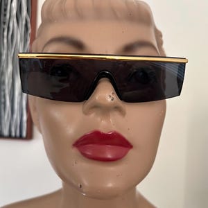 Puede incluir: Gafas de sol negras con una barra superior dorada. Las gafas de sol tienen forma rectangular y lentes oscuras. Las gafas de sol se exhiben en una cabeza de maniquí con lápiz labial rojo.