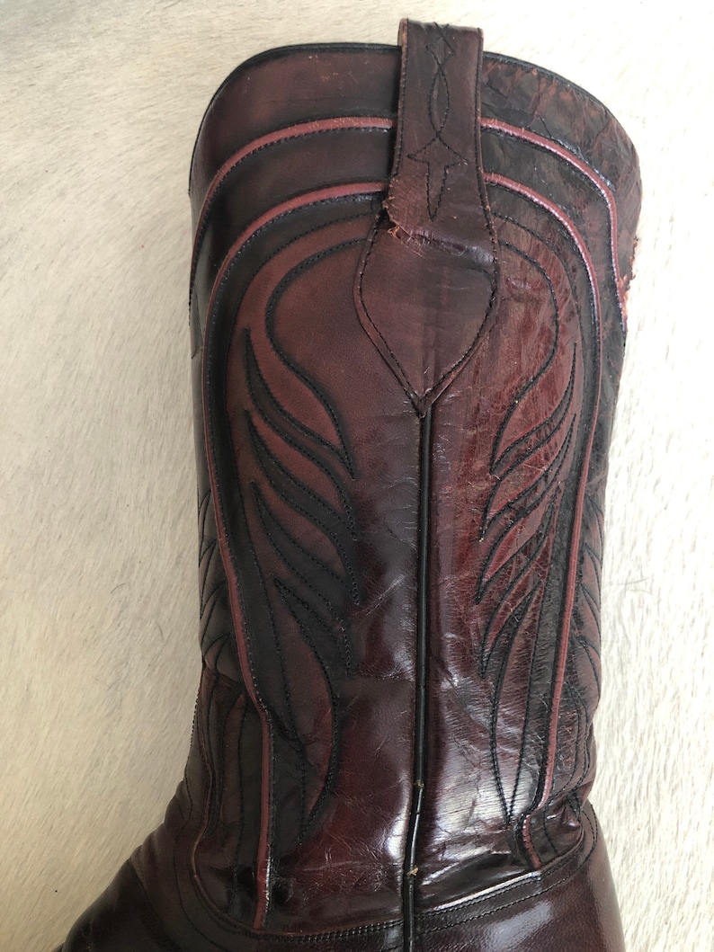 Puede incluir: Primer plano de una bota de vaquero de cuero marr&oacute;n. La bota presenta costuras decorativas negras y rojas, una puntera puntiaguda y una ca&ntilde;a alta. El cuero tiene un acabado brillante.