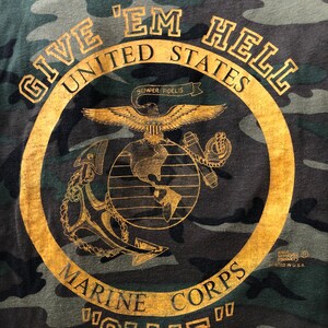 Op de afbeelding: T-shirt met camouflagepatroon en een goudkleurig embleem. Het embleem toont een adelaar, anker, wereldbol en de tekst "GIVE 'EM HELL UNITED STATES MARINE CORPS" en "OLLIE". Het shirt is bedrukt in de VS.