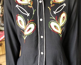 May include: Black long-sleeve button-up shirt with white trim and colorful embroidered floral designs. The shirt features a pointed collar and decorative stitching around the chest. The cuffs and collar are trimmed with white piping.