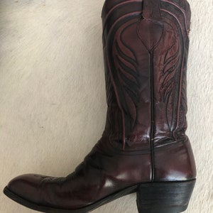 Puede incluir: Bota de vaquero de cuero burdeos con costuras decorativas. La bota presenta una puntera puntiaguda, un tac&oacute;n apilado y una ca&ntilde;a alta con patrones intrincados. El cuero tiene un acabado brillante y la bota parece desgastada.
