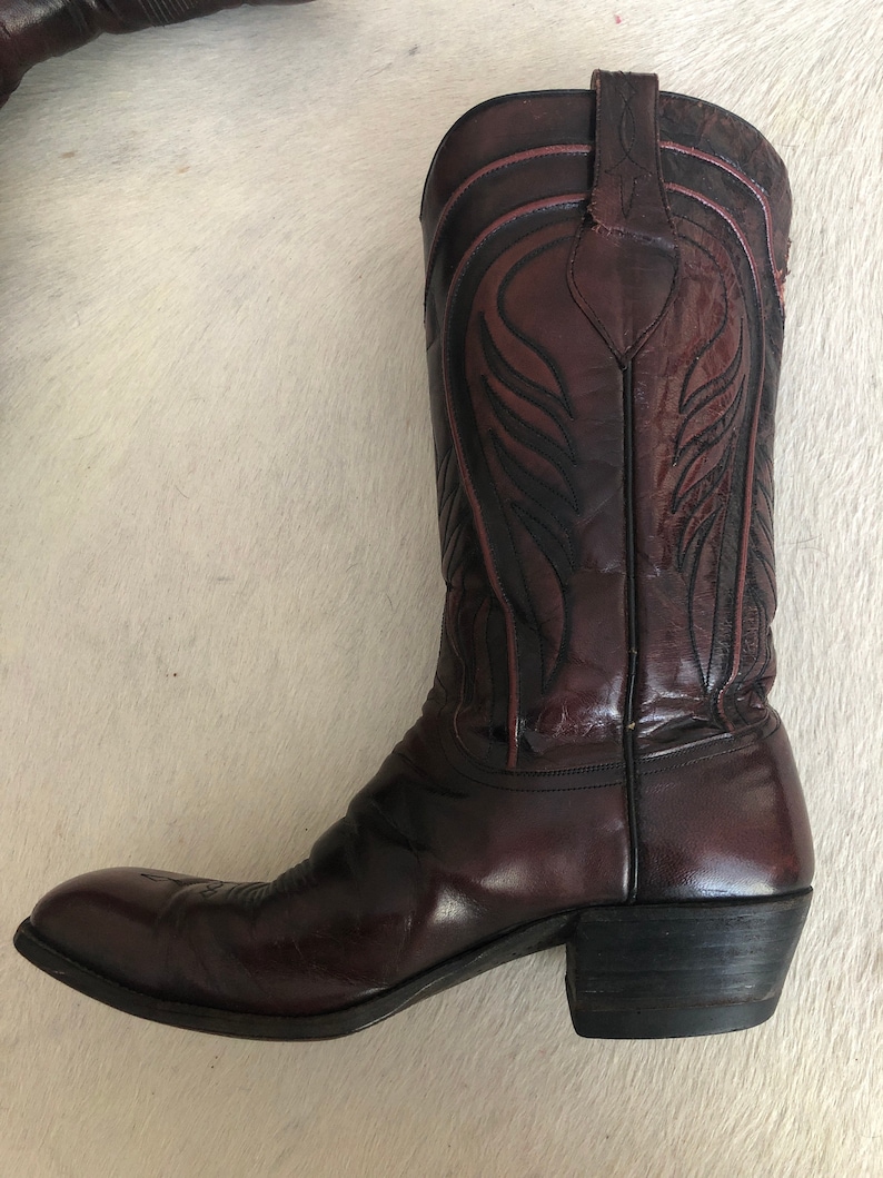 Puede incluir: Bota de vaquero de cuero burdeos con costuras decorativas. La bota presenta una puntera puntiaguda, un tac&oacute;n apilado y una ca&ntilde;a alta con patrones intrincados. El cuero tiene un acabado brillante y la bota parece desgastada.
