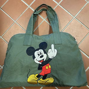 Könnte beinhalten: Olivgrüne Tragetasche mit einem gestickten Bild von Micky Maus, die den Mittelfinger zeigt. Die Tasche hat zwei Griffe und einen Reißverschluss. Micky Maus trägt rote Shorts und gelbe Schuhe.