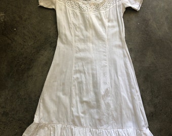 May include: A white dress with a square neckline and short sleeves. The dress has a detailed lace yoke and a ruffled hem with floral cutouts. The dress is made of a lightweight fabric, suitable for warmer weather.