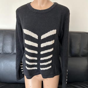 May include: Black sweater with a white rib cage design. The long-sleeved sweater has a crew neck and white stripes on the sleeves. The sweater is displayed on a mannequin.