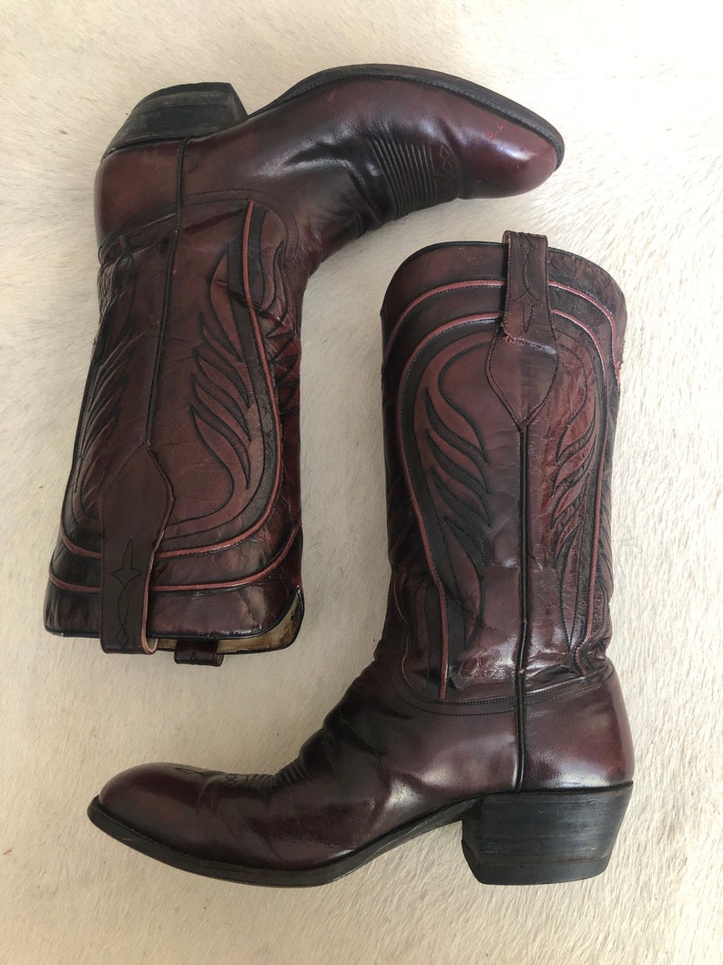 Puede incluir: Un par de botas de vaquero burdeos con costuras decorativas. Las botas tienen una punta puntiaguda, un tac&oacute;n apilado y un dise&ntilde;o sin cordones. El cuero est&aacute; pulido y tiene un acabado brillante. Las botas son de estilo occidental cl&aacute;sico.