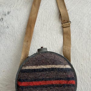 May include: A circular vintage canteen with a striped wool cover in brown, red, and white. It features a metal rim and cap, and a tan canvas strap. The canteen is designed to be carried and shows signs of wear.