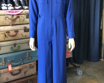 Puede incluir: Mono azul vibrante con cuello camisero y mangas largas. La prenda de una pieza presenta una cremallera frontal, dos bolsillos en el pecho con cierre de botón y pantalones de pernera ancha. El mono se exhibe en un maniquí.