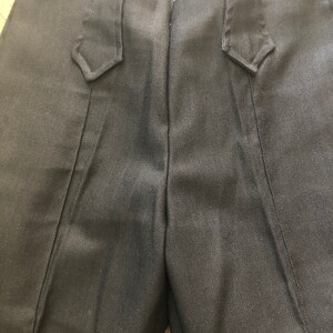 May include: Close-up of black pants with a tailored design. The pants feature a high waist and decorative details near the pockets. The fabric has a subtle sheen, and the overall style suggests formal or semi-formal wear.