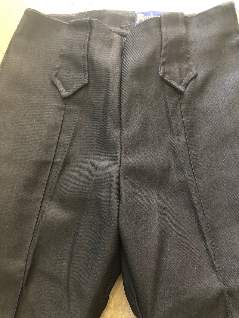 May include: Close-up of black pants with a tailored design. The pants feature a high waist and decorative details near the pockets. The fabric has a subtle sheen, and the overall style suggests formal or semi-formal wear.