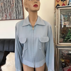 May include: A light blue jacket with a collared neckline, two front pockets, and a gold zipper closure. The jacket is displayed on a mannequin. The jacket's sleeves are long and the fabric appears to be lightweight.