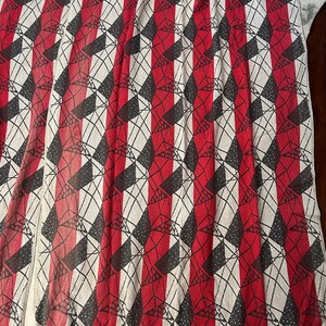 May include: A patterned textile with a repeating geometric design. Vertical red stripes alternate with white sections. Black diamond shapes and crisscrossing lines create a textured effect. The fabric is draped, showing its folds and texture.