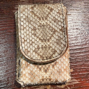 May include: A rectangular case with a snakeskin texture in shades of beige and brown, featuring a flap closure. The edges are stitched with black thread. The case is designed to hold small items.