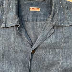 May include: A blue linen shirt with a collar and button-down front. The shirt has a pocket on the lower right side and a label that reads "KAPITAL MADE IN JAPAN". The shirt has a relaxed fit and is made of a lightweight fabric.