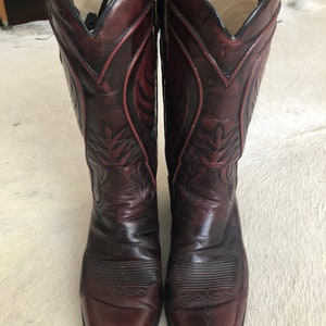 Puede incluir: Un par de botas de vaquero burdeos. Las botas tienen una puntera puntiaguda y costuras decorativas a lo largo de la ca&ntilde;a. El cuero es de color burdeos oscuro con detalles m&aacute;s oscuros. El interior es de color beige claro.