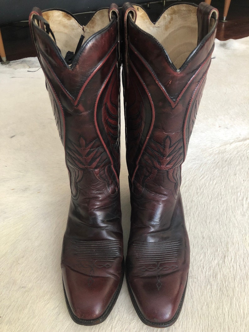Puede incluir: Un par de botas de vaquero burdeos. Las botas tienen una puntera puntiaguda y costuras decorativas a lo largo de la ca&ntilde;a. El cuero es de color burdeos oscuro con detalles m&aacute;s oscuros. El interior es de color beige claro.