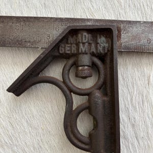 May include: A dark brown, cast iron square tool with a textured finish, featuring the embossed text "MADE IN GERMANY". The tool is positioned against a metal ruler, showing the base edge aligns with the 18 cm mark on the metric scale.