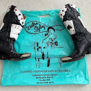 May include: A pair of black leather cowboy boots with white and red playing card designs on the shafts. The boots have silver-toned metal toe caps and are displayed on a turquoise shopping bag with the text "Western Styling".