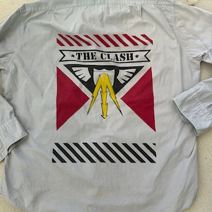 Puede incluir: Camisa abotonada azul claro con un gráfico en la espalda. El gráfico presenta el texto "THE CLASH" sobre un diseño rojo y negro con un rayo amarillo.