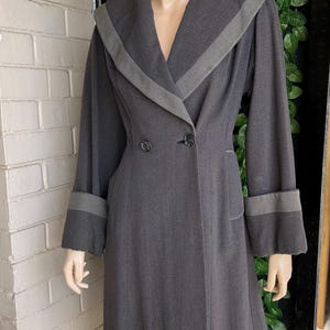 Könnte beinhalten: Ein vintage Mantel in Anthrazitgrau mit einem kontrastierenden grauen Kragen und Manschetten. Der Mantel hat ein doppelreihiges Design mit zwei dekorativen Knöpfen und einer taillierten Passform. Der Mantel wird auf einer Schaufensterpuppe präsentiert.