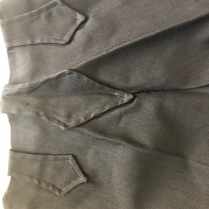 May include: Close-up of a black garment with three decorative, diamond-shaped flaps. The fabric appears to be a smooth, woven material. The flaps are sewn onto the garment, adding a tailored detail. The overall aesthetic is simple and elegant.