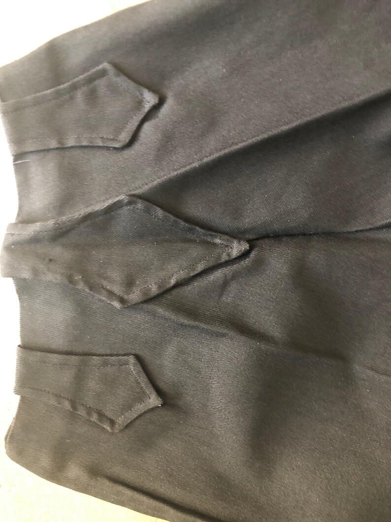 May include: Close-up of a black garment with three decorative, diamond-shaped flaps. The fabric appears to be a smooth, woven material. The flaps are sewn onto the garment, adding a tailored detail. The overall aesthetic is simple and elegant.