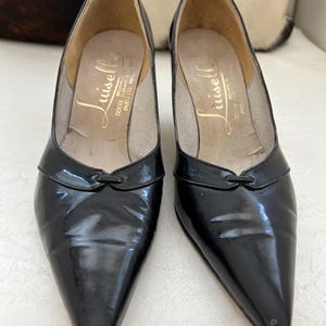 May include: A pair of black patent leather pumps with pointed toes. The shoes have a small bow detail on the vamp and a low heel. The insoles are tan with gold lettering. The shoes are vintage and appear to be unworn.