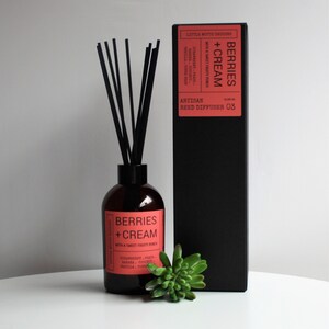 May include: A black box with a red label that reads "BERRIES + CREAM" with a sweet fruity punch. The box contains a brown glass bottle with black reeds and a red label that reads "BERRIES + CREAM" with a sweet fruity punch. The bottle is sitting on a white surface.