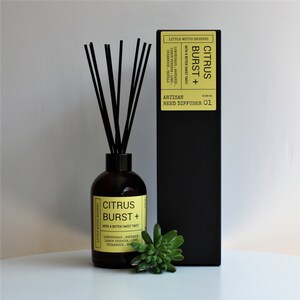 May include: A black and yellow box with the text "Citrus Burst +" and "Artisan Reed Diffuser 01". A brown glass bottle with a black lid and black reeds is in front of the box. The bottle has a yellow label with the text "Citrus Burst +" and a list of ingredients.