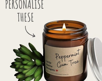 Op de afbeelding: Een bruine glazen kaars pot met een witte kaars erin. De pot heeft een label met de tekst "Peppermint Gum Tree" en "Little Motto Designs." De kaars is aangestoken en de vlam is zichtbaar.