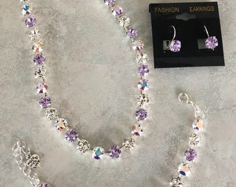 May include: A silver necklace, bracelet, and earrings set with clear and light purple crystals. The jewelry is on a gray background. The earrings are labeled "Fashion Earrings".