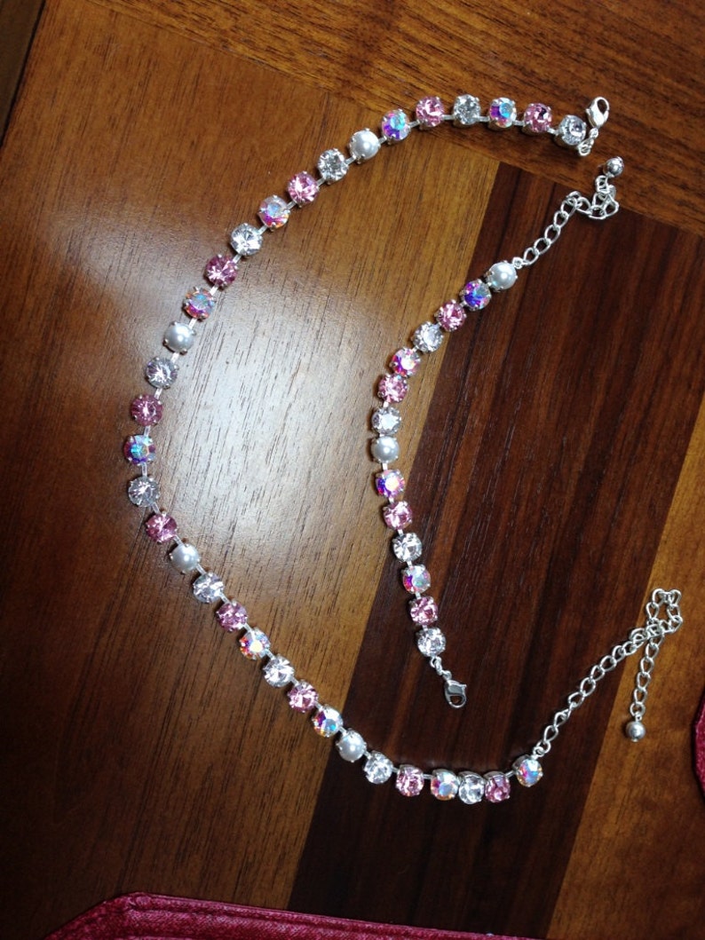 May include: A silver-toned necklace with a delicate design. The necklace features alternating pink and clear crystals, with iridescent and white pearl accents. The necklace is displayed on a wooden surface.