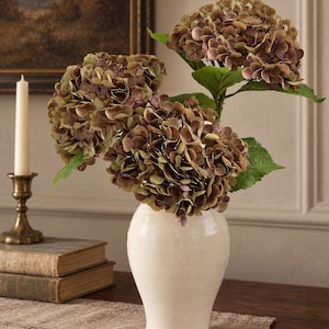 May include: A bouquet of artificial hydrangea flowers with muted brown, green, and purple petals arranged in a smooth, off-white ceramic vase. The flowers feature dense, rounded clusters of small blossoms and several green leaves on stems.