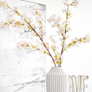 Peut inclure: Vase en céramique blanche avec un design texturé et côtelé, contenant un bouquet de fleurs de cerisier blanches artificielles avec des tiges vertes.