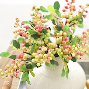 Może przedstawiać: Biały ceramiczny wazon z fakturowaną powierzchnią zawiera bukiet sztucznych gałązek jagód. Jagody są mieszanką różowych i zielonych.