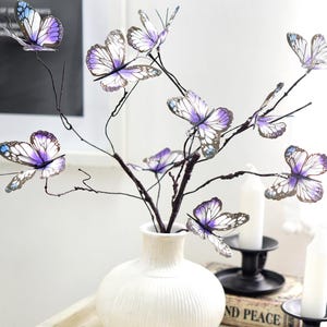 以下が含まれることがあります： 白いセラミックの花瓶には、蝶の枝の装飾的なアレンジメントが入っています。 蝶は紫と白の羽と黒のアクセントを持っています。 枝は濃い茶色で上に伸びています。 花瓶は白いトレイの上にあります。