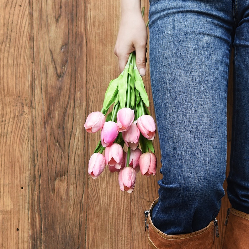 K&ouml;nnte beinhalten: Ein Strau&szlig; rosa Tulpen, gehalten von einer Person in blauen Jeans und braunen Stiefeln.