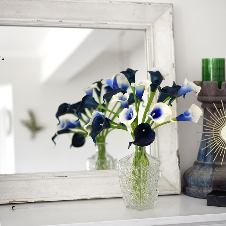 May include: A bouquet of white and blue calla lilies arranged in a clear glass vase with a textured design. The vase is sitting on a white surface in front of a white framed mirror.