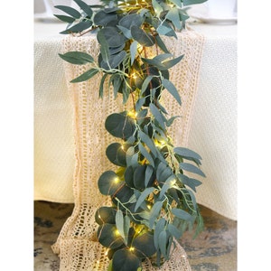 Op de afbeelding: Een eucalyptus slinger met ronde en langwerpige groene bladeren, verlicht door kleine, warmwitte feeërieke lichtjes. De slinger is gedrapeerd over een crèmekleurige tafelloper, wat een zachte, natuurlijke uitstraling creëert.