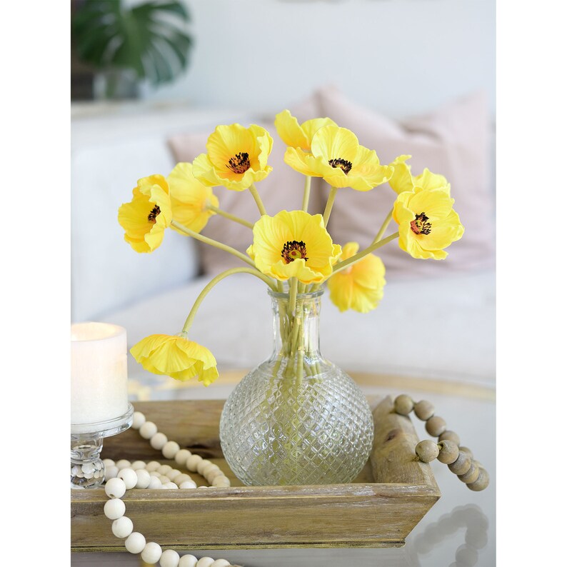 Puede incluir: Un ramo de flores de amapola de seda amarilla en un jarr&oacute;n de vidrio transparente con una superficie texturizada. El jarr&oacute;n est&aacute; sentado en una bandeja de madera con cuentas blancas.