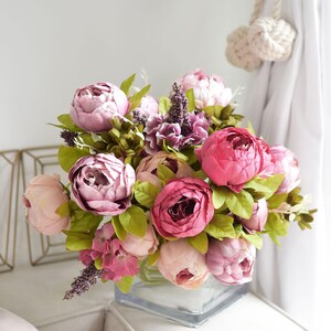 Puede incluir: Un ramo de peon&iacute;as artificiales de color rosa y morado en un jarr&oacute;n de vidrio transparente. Las flores est&aacute;n dispuestas en un estilo suelto y natural.