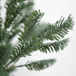May include: Close-up of a green artificial pine branch with needle-like leaves. The branch has a frosted appearance, with a light dusting of white. The background is a plain white, highlighting the details of the evergreen foliage.
