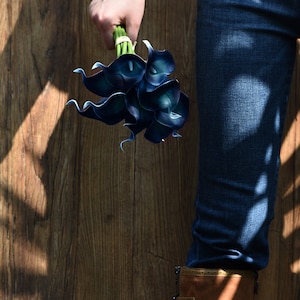May include: A bouquet of dark blue calla lilies held by a person wearing blue jeans and brown boots.