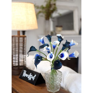 May include: A bouquet of white and blue calla lilies in a clear glass vase. The vase has a textured surface. The flowers are arranged in a vase on a wooden table.