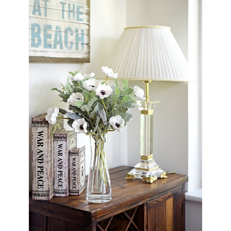 May include: A white vase with a bouquet of white flowers with dark centers. The vase is on a wooden table with three books stacked on the left side of the vase. The books are titled "War and Peace". A white lamp with a pleated shade is on the right side of the vase.