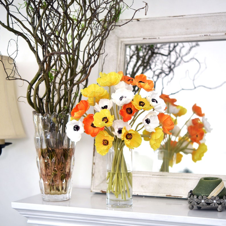 Puede incluir: Un ramo de flores amarillas, blancas y naranjas en un jarr&oacute;n de vidrio transparente. Las flores est&aacute;n dispuestas en un jarr&oacute;n sobre una repisa blanca frente a un espejo blanco.